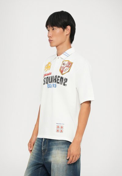 Cămașă polo albă cu mânecă scurtă, cu patches brodate, guler în dungi albastre și text grafic. Poartă cu blugi albaștri decolorați.