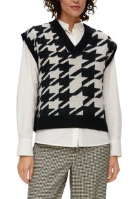 Gilet sans manches en tricot avec un motif pied-de-poule noir et blanc, doté d'un décolleté en V profond et d'un ourlet côtelé. Porté par-dessus une chemise blanche.