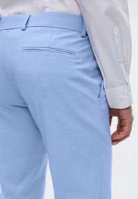 Calças ajustadas azul claro com uma textura suave, apresentando um bolso traseiro e passantes para cinto, combinadas com uma camisa branca de mangas longas com botões.
