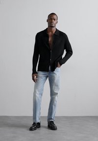 Svart ribbad stickad cardigan, knäppt, bärs över en bar bröstkorg. Ljusblå rakbenade jeans och svarta klumpiga skor fullbordar stilen.