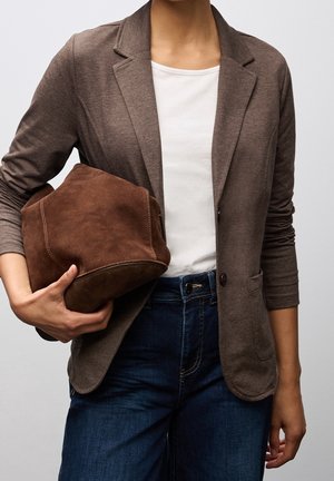 Persoon draagt een bruine blazer en wit overhemd, houdt een zachte bruine suède tas vast, met blauwe spijkerbroek zichtbaar vanaf de taille naar beneden.