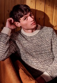 Pulover tricotat cu un model texturat în nuanțe de maro și crem, având un guler decorat cu coaste de crem. Modelul pozează într-o manieră casual pe o canapea maro.