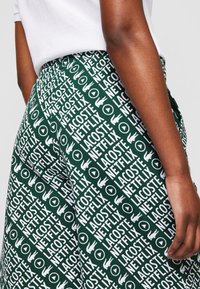 Personne portant un short vert avec des motifs de texte blancs répétitifs "Lacoste" et "Netflix" et le logo crocodile Lacoste.