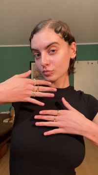 Zlaté prsteny v různých designech, včetně hladkých kroužků a texturovaných detailů, nošené na prstech; v pozadí je tmavá zeď a světlý nábytek.