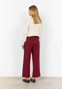 Frau steht mit dem Rücken zur Kamera, trägt einen cremefarbenen Pullover, hoch geschnittene weite bordeauxrote Hose und dunkle, mit Nieten verzierte flache Schuhe auf einem grauen Boden.