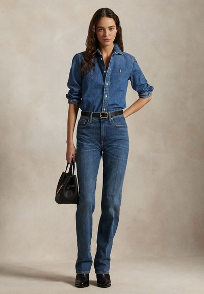 Moteris vilki džinsinę marškinius ir džinsus su juodu diržu, laiko juodą rankinę, stovi prieš neutralią smėlio spalvos foną.