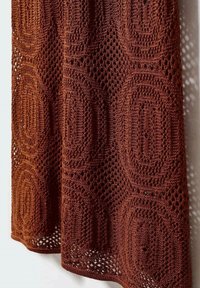 Tissu en crochet brun avec des motifs hexagonaux et des détails en maille ouverte, drapé contre un fond clair.