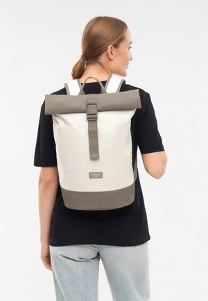 Ung person med lyst brunt hår i hestehale iført sort skjorte og lyse jeans, bærende på en beige og grå roll-top rygsæk.