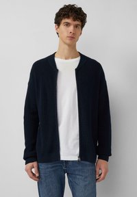 Marineblauer, gerippter Strick-Cardigan mit abgerundetem Kragen, durchgehendem Reißverschluss und langen Ärmeln. Kombiniert mit einem weißen T-Shirt und blauen Jeans.