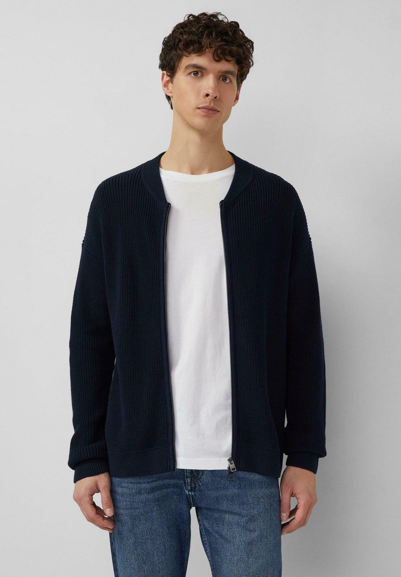 Marineblauer, gerippter Strick-Cardigan mit abgerundetem Kragen, durchgehendem Reißverschluss und langen Ärmeln. Kombiniert mit einem weißen T-Shirt und blauen Jeans.