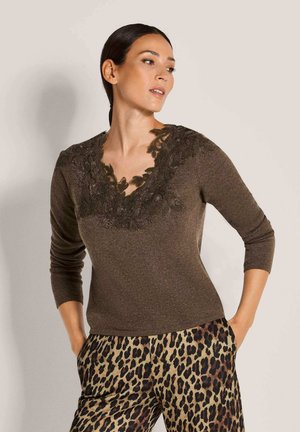 Vrouw met donker haar draagt een bruine top met lange mouwen met een bloemenkant halslijn en een broek met luipaardprint, staand met handen in de zakken.