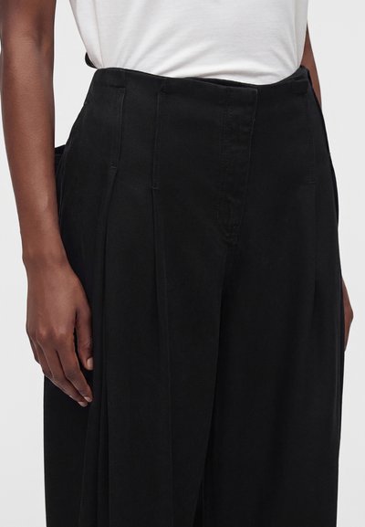Gros plan d’une personne portant un pantalon noir taille haute à jambes larges et une chemise blanche rentrée, montrant une main gauche détendue sur le côté.