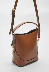 Sac seau en cuir brun au design structuré, avec des accessoires dorés, une bandoulière réglable et des coutures contrastées le long des bords.