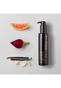Svart flaska med pump, märkt "essens absolut", omgiven av grapefruktskiva, rosknopp, vaniljstänger och vita blommor på en grå bakgrund.