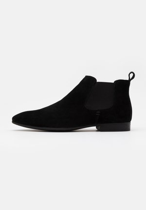 Les bottines Chelsea en daim noir présentent des panneaux latéraux élastiques, une boucle d'aisance et un talon bas et carré, avec une texture lisse et une silhouette épurée.