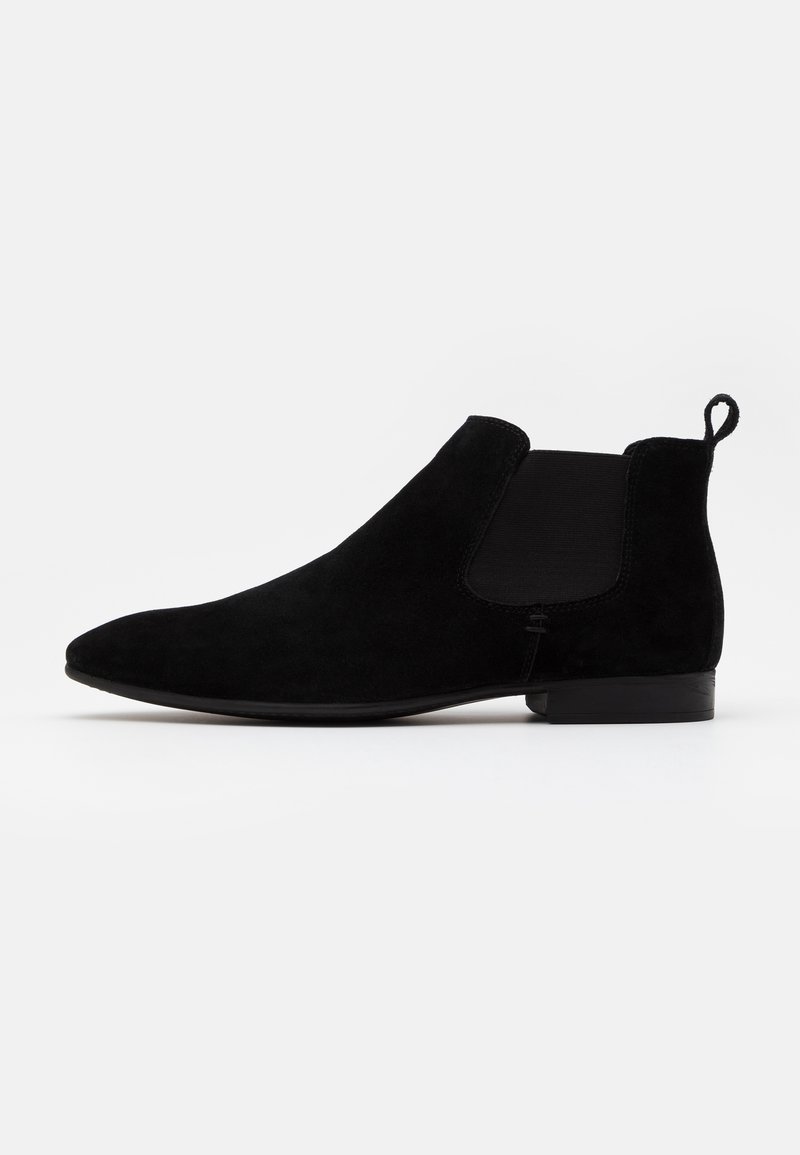 Les bottines Chelsea en daim noir présentent des panneaux latéraux élastiques, une boucle d'aisance et un talon bas et carré, avec une texture lisse et une silhouette épurée.