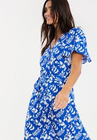 Blå klänning med vita bladmönster, korta svävande ärmar, V-ringning och knytning i midjan. Tillverkad av lätt tyg, lämplig för varma väderförhållanden.