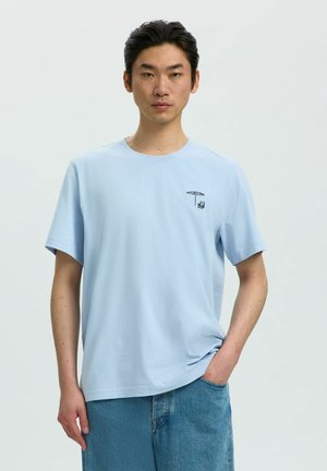 Jeune homme portant un t-shirt bleu clair avec un petit motif noir d'un parapluie et d'une chaise, associé à un jean en denim bleu, sur un fond uni.