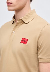 Άνδρας που φοράει μπεζ πόλο μπλουζάκι με ριμπ ύφασμα στο γιακά και τα μανίκια, με ένα μικρό κόκκινο τετράγωνο πατς με την επιγραφή "HUGO" στο στήθος.