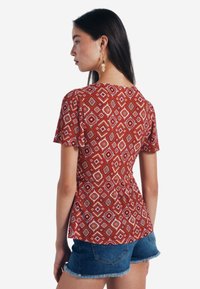 Rotes gemustertes Oberteil mit kurzen Ärmeln, Diamant- und Blumendesigns, kombiniert mit Jeansshorts. Weicher Stoff mit Peplum-Silhouette.
