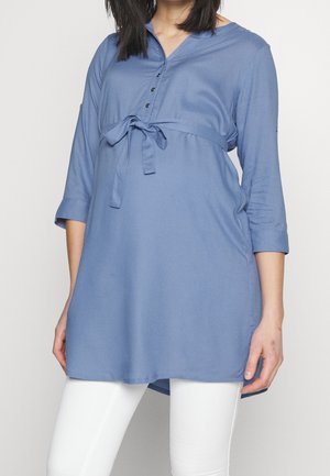 Vrouw die een losse blauwe tuniek draagt met een strik in de taille, driekwartmouwen en een witte broek.