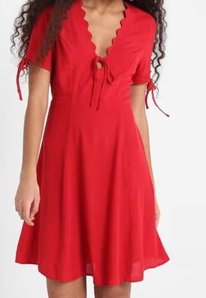 Rød knælængde kjole med bølget udskæring, korte ærmer bundet med sløjfer og en tætsiddende talje, der går ud i et flagrende skørt.