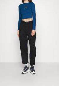 Modrý dlouhý rukáv cropped top s výřezem a zeleným logem; černé cargo kalhoty s kapsami; bílé a černé sneakers s modrými akcenty.