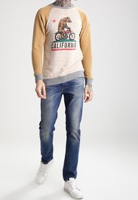 Grå och senapsgul långärmad sweatshirt med en grafisk bild av en björn från Kalifornien, kombinerad med mörkblå jeans och vita sneakers.