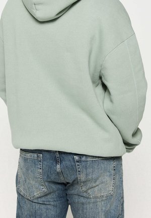 Felpa verde con cappuccio e fit rilassato, caratterizzata da polsini e orlo a costine, abbinata a jeans blu scoloriti con tasche posteriori.