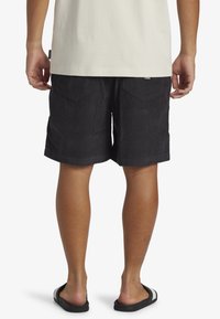 Svarte corduroy shorts med en afslappet pasform, synlig syning og to baglommer. Bæres med sorte slide sandaler, set bagfra.
