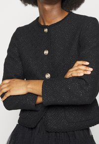 Černý tvídový blejzr s texturovaným povrchem, vybavený třemi zlatými kulatými knoflíky, kulatým výstřihem a zkráceným střihem s kapsami.