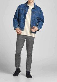 Giacca di jeans con tasche sul petto sopra una camicia a righe bianche e gialle, abbinata a pantaloni grigi slim-fit e scarpe scure.