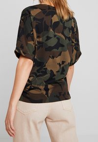 Kamouflage T-shirt i nyanser av grönt, brunt och svart; korta ärmar, avslappnad passform, tillverkad av bomull, med en subtil logodetalj på nederkanten.