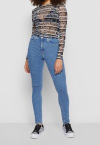 Modré skinny džíny s vysokým pasem a hladkým povrchem, spárované s průhledným černým topem s dlouhým rukávem, na kterém se opakovan�ě objevuje značka Calvin Klein.