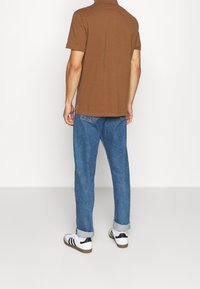 Brązowa koszulka polo z krótkim rękawem, noszona z niebieskimi dżinsami i białymi trampkami z czarnymi paskami. Widoczne podwinięte mankiety na dżinsach.