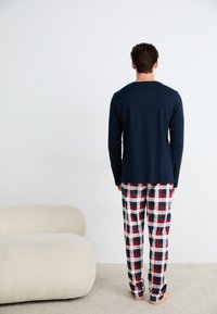 Camicia a maniche lunghe blu navy abbinata a pantaloni da pigiama a quadri rossi, bianchi e blu; tessuto morbido e vestibilità rilassata; in piedi a piedi nudi.