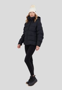 Černá péřová bunda s prošívanými sekcemi, zapínání na zip a kapucí; nošena přes černé skinny džíny a černé šněrovací boty; krémová pletená čepice.