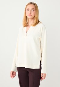 Haut à manches longues couleur crème avec un col en V et des fentes latérales, fabriqué en tissu doux, associé à un pantalon marron foncé.