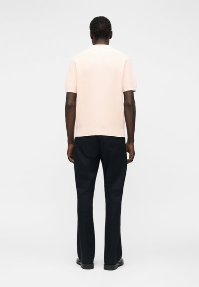 Άνδρας που στέκεται με την πλάτη προς τα εμπρός φορώντας ένα ανοιχτό ροζ κοντομάνικο πλουαζέ πόλο με υφή, μαύρο παντελόνι ίσιας γραμμής και μαύρα παπούτσια σε λευκό φόντο.