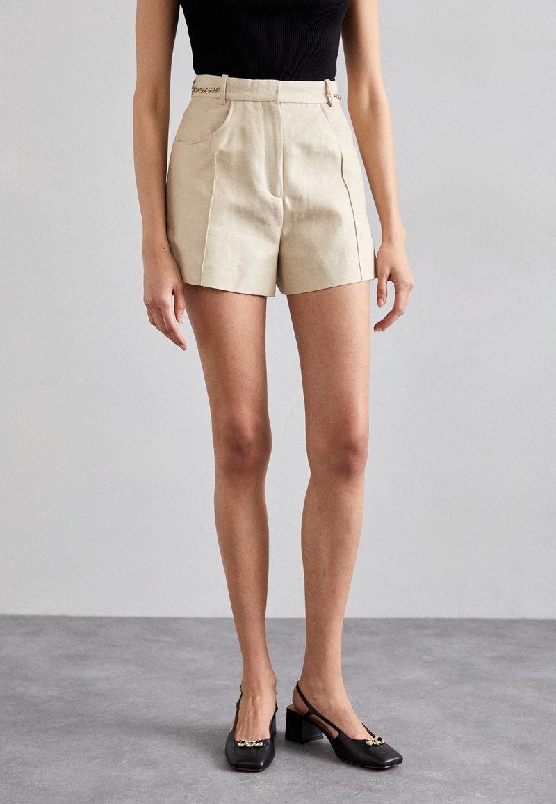 Femme portant un short beige taille haute avec poches latérales, un top sans manches noir et des chaussures slingback noires à talon bas avec un détail doré.