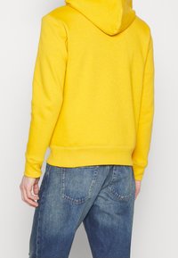 Sudadera amarilla con capucha y textura suave, con un bolsillo tipo canguro, combinada con jeans de mezclilla azul que tienen dos bolsillos traseros.