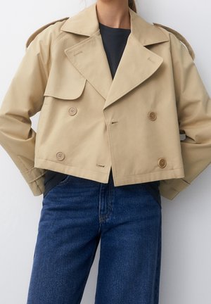 Trench court beige à double boutonnage porté sur un haut noir et un jean bleu, mains dans les poches.