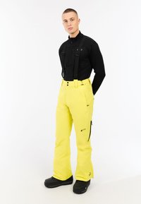 Geel skibroek met verstelbare bretels, sluiting met voorste knoop, zijritsen en logodetail. Draag het met een zwart zip-up bovenstuk en zwarte laarzen.