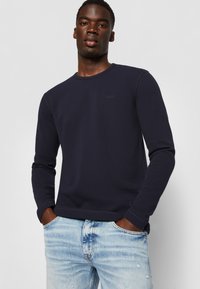 Marineblaues Langarm-T-Shirt mit gerippter Textur und Rundhalsausschnitt, mit einem dezenten Logo auf der Brust, kombiniert mit hellblauen, ausgewaschenen Jeans.
