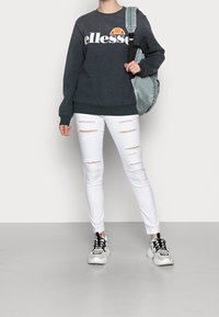 Felpa grigia con logo "ellesse", jeans skinny bianchi strappati e sneakers chunky in bianco e nero. Il modello porta uno zaino verde chiaro.