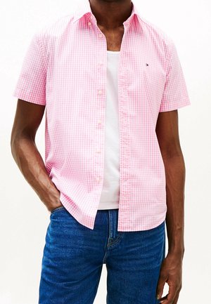 Mann trägt ein kurzärmeliges, rosa-weiß kariertes Hemd über einem weißen T-Shirt und blauen Jeans, eine Hand in der Tasche.