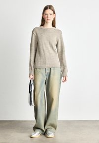 Light brown knitted sweater with textured detailing on sleeves, paired with loose light blue denim jeans and silver sneakers. Holding a metallic bag.