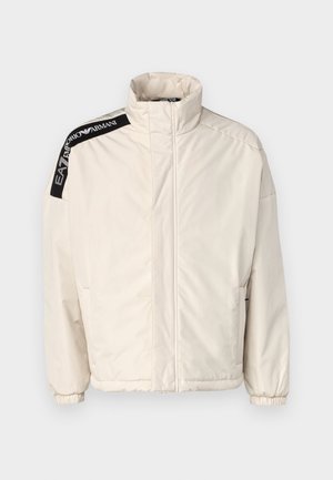 Polstret lys beige jakke med høy krage. Har svarte logoaksenter på skulderen og elastiske mansjetter for tettsittende bruk.