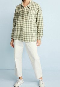 Green and white checkered shirt jacket with two front pockets and button closure, paired with white pants and light sneakers.