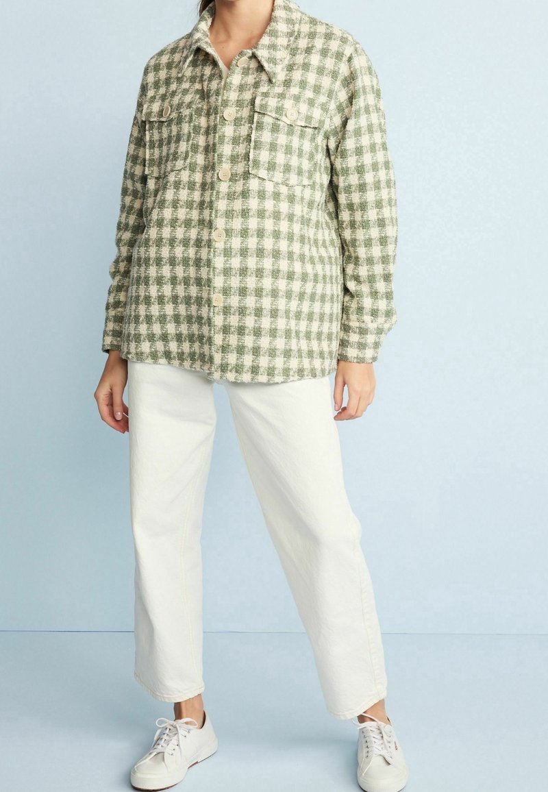 Green and white checkered shirt jacket with two front pockets and button closure, paired with white pants and light sneakers.